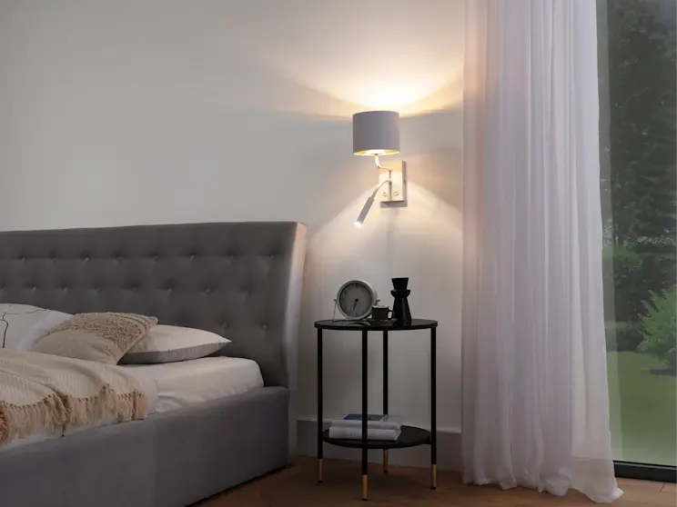 Przytulny kącik przy łóżku Milano: szare wezgłowie, czarny stolik nocny i kinkiet Vespillo z abażurem oraz lampką do czytania.