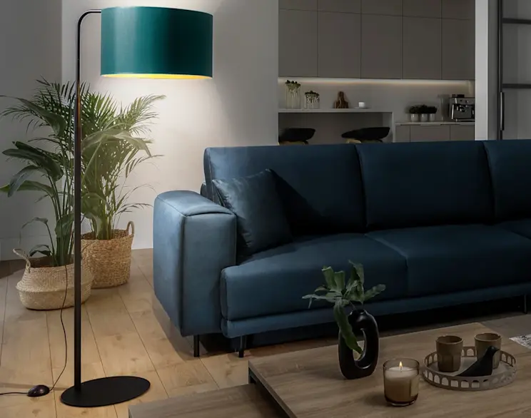 Lampa stojąca podłogowa Vespillo z zielonym abażurem i czarną podstawą ustawiona obok granatowej sofy. W tle widać rośliny w plecionych koszach oraz nowoczesną kuchnię, a ciepłe światło lampy buduje przytulny klimat salonu.