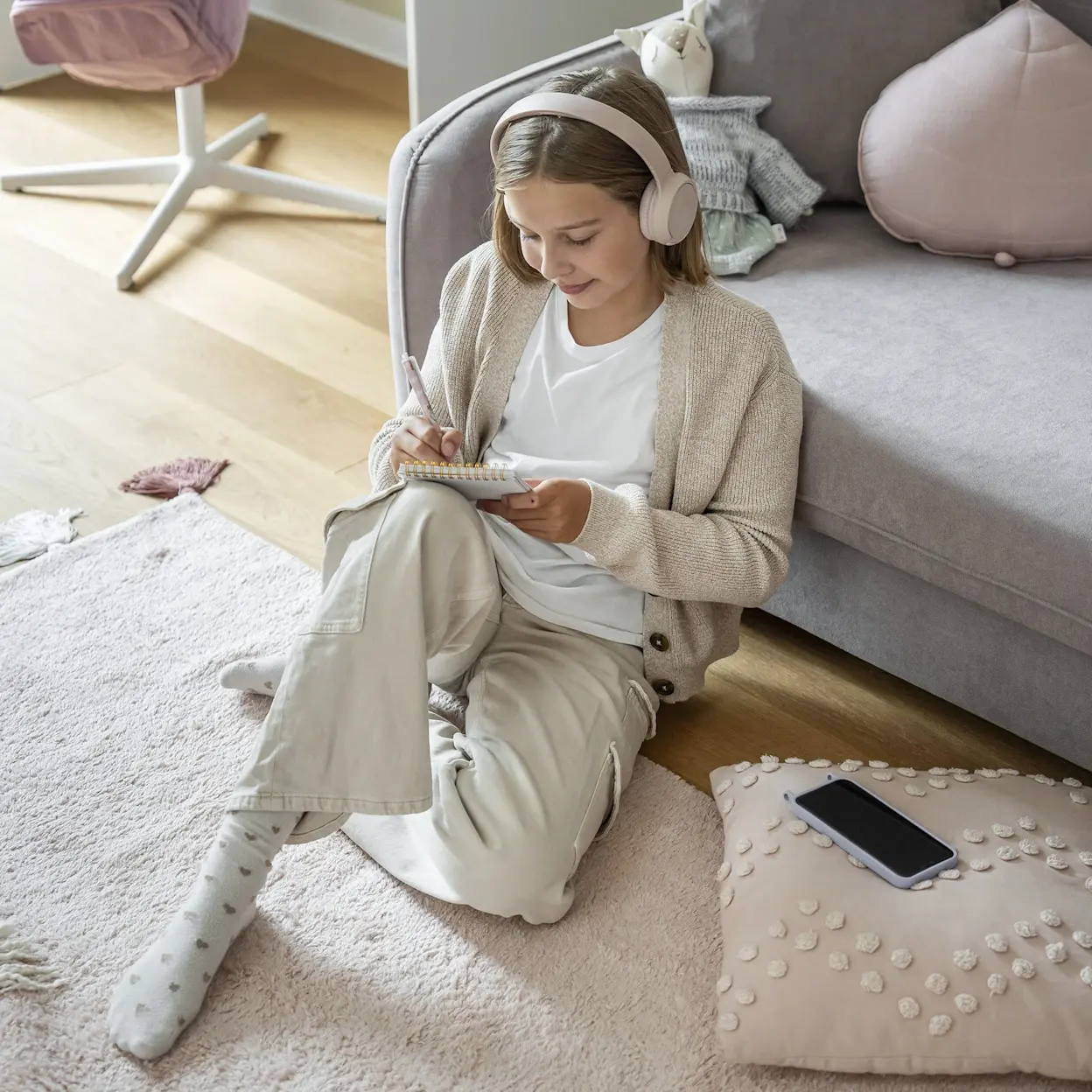 Dziewczynka w słuchawkach siedzi na miękkim dywanie przy szarej kanapie Laine w pokoju dziecięcym i robi notatki. W tle widać biurko Nevy, pastelowe dodatki oraz poduszki i maskotkę na sofie.