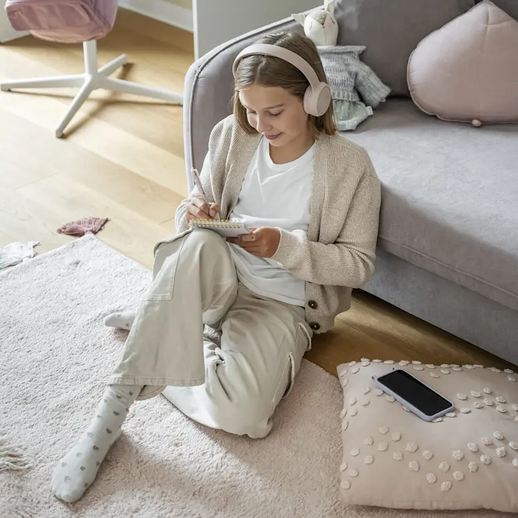 Dziewczynka w słuchawkach siedzi na miękkim dywanie przy szarej kanapie Laine w pokoju dziecięcym i robi notatki. W tle widać biurko Nevy, pastelowe dodatki oraz poduszki i maskotkę na sofie.
