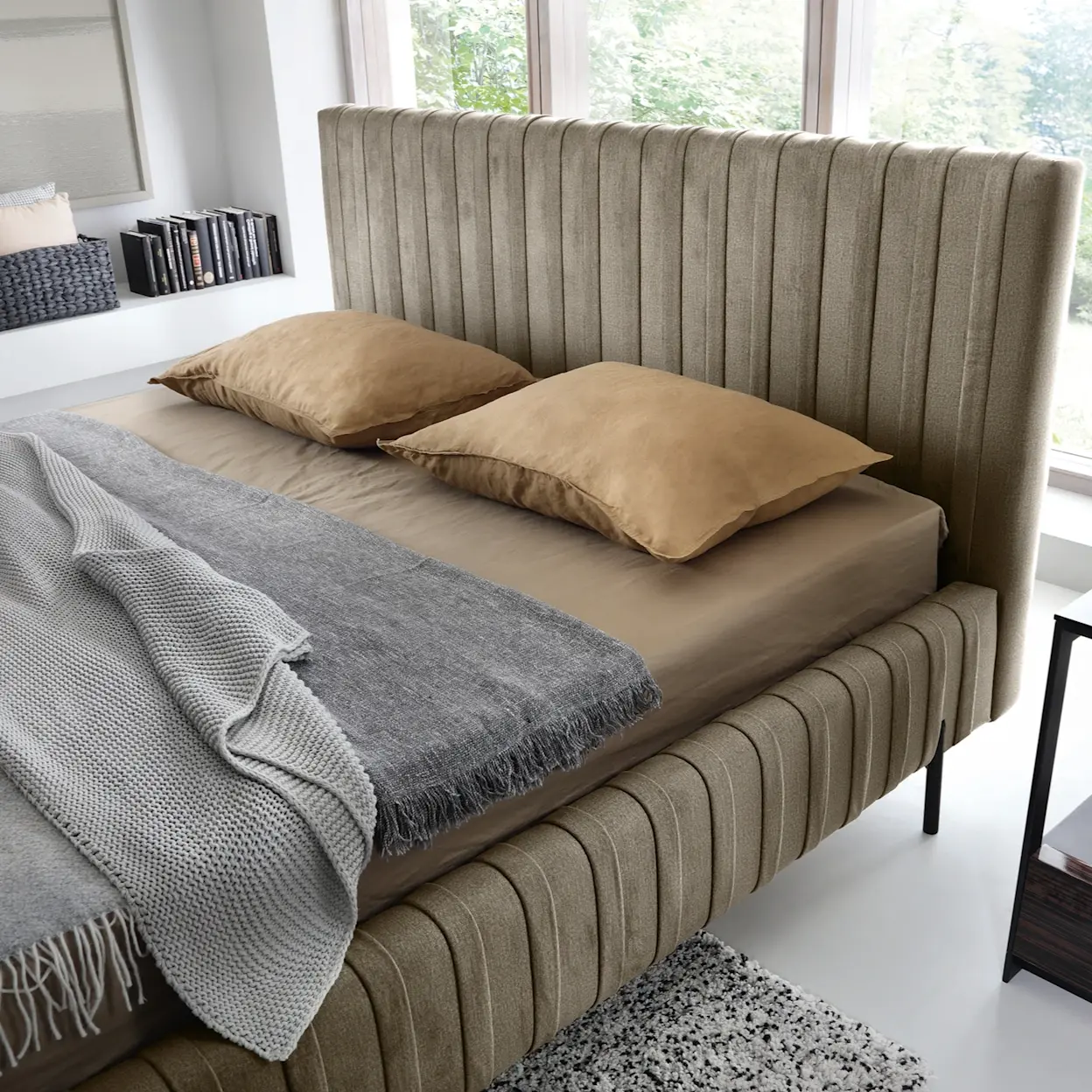 Komfortowe łóżko Plissa Nube 20 — tapicerowana rama i wysokie wezgłowie w pionowe przeszycia, beżowe prześcieradło, szary pled i ciepłe poduszki w jasnej, naturalnej sypialni.