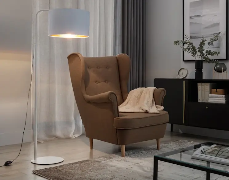 Lampa stojąca Vespillo w kolorze białym ze złotym wnętrzem abażuru oświetla kącik wypoczynkowy z brązowym fotelem typu uszak. W tle szare zasłony i czarna komoda z dekoracjami, a na pierwszym planie widać fragment stolika i dywanu.