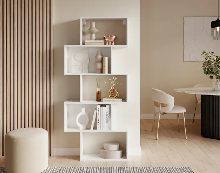 Biały regał schodkowy Sottis ustawiony przy lamelach w jasnym, beżowym wnętrzu. Na półkach książki i dekoracje, a obok stoi pufa Linsy w tkaninie Monolith 09, tworząc spójną aranżację salonu.