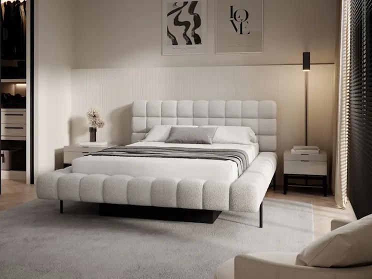 Jasna sypialnia z łóżkiem Magentta: duży, tapicerowany model z pikowaniami, białą pościelą i szarą narzutą. Obok znajdują się minimalistyczne stoliki nocne, dekoracje oraz ciepłe oświetlenie budujące przytulny klimat.