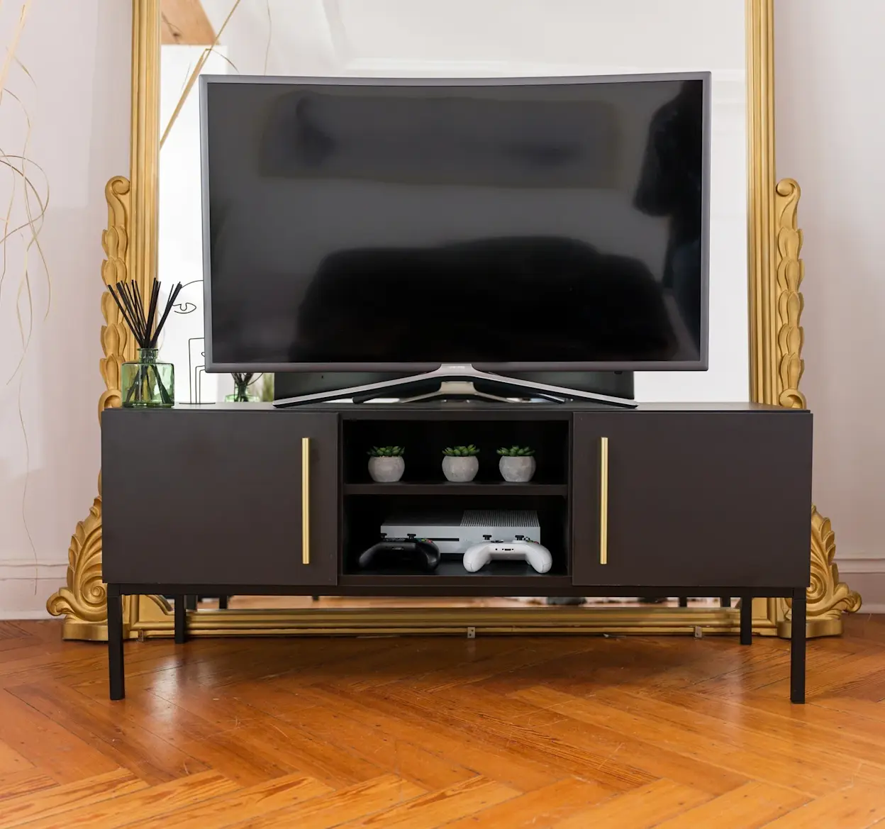 Czarna szafka RTV Lanzzi z eleganckimi złotymi uchwytami, ustawiona przed dużym lustrem, z telewizorem i dekoracjami na drewnianej podłodze.