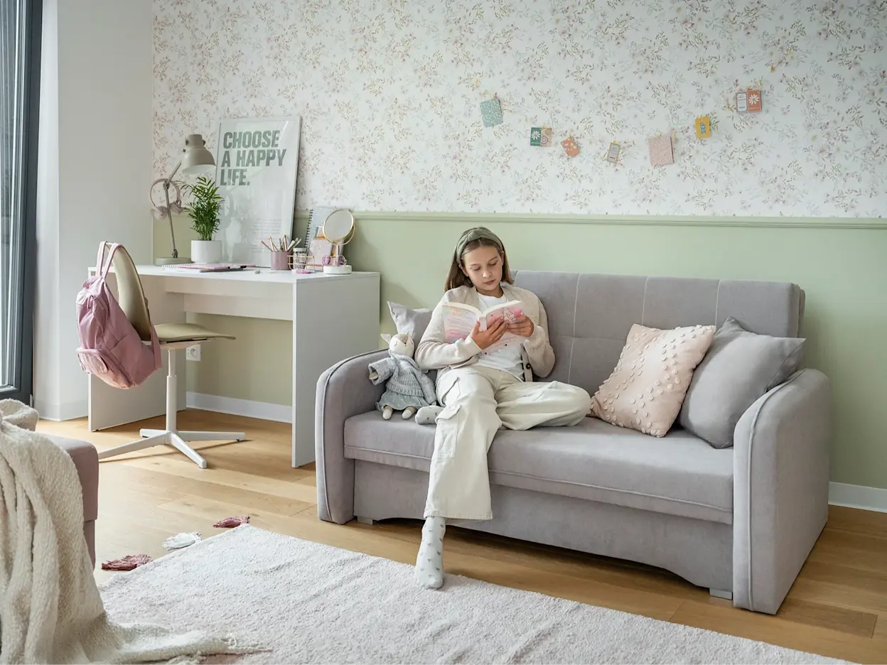 Kanapa Laine obok biurka Nevy w jasnym, młodzieżowym wnętrzu; dziewczynka siedzi na sofie i czyta, otoczona miękkimi tekstyliami i dekoracjami.