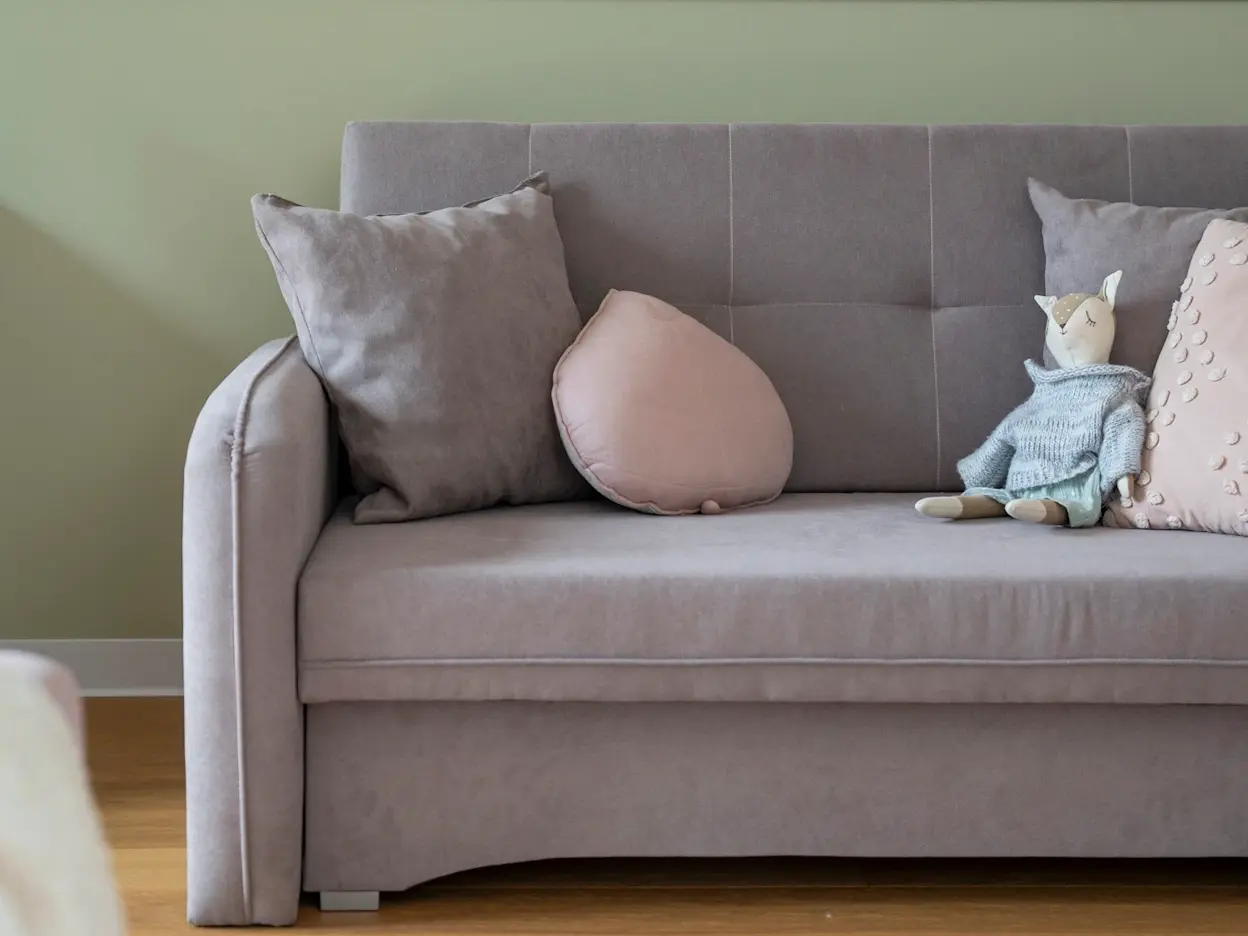 Kanapa Laine w dziecięcej aranżacji: miękka, szara sofa z prostym oparciem i zestawem poduszek w pastelach. Dekoracyjna tapeta w kwiaty i zielona ściana tworzą przytulne tło do odpoczynku i zabawy.