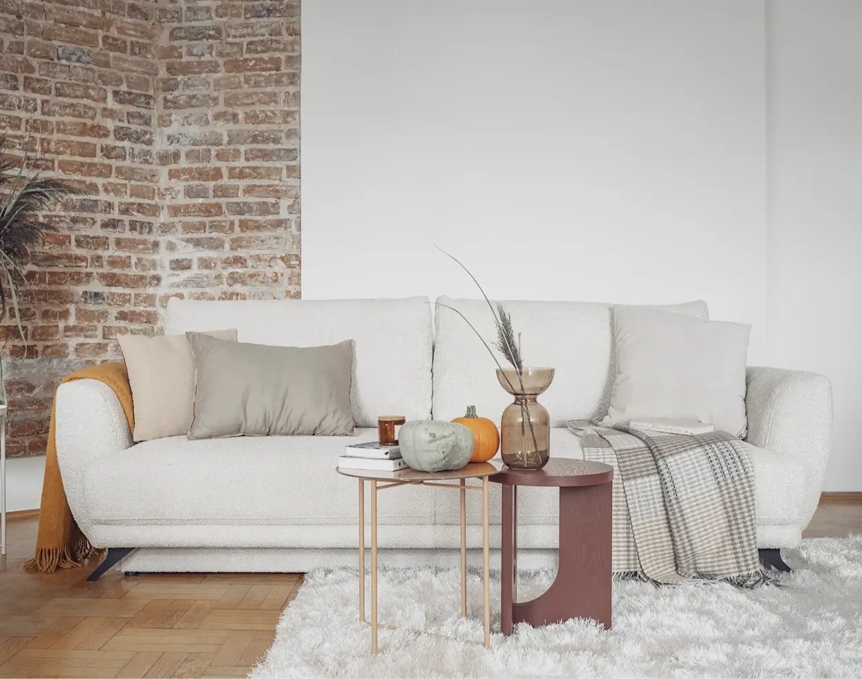 Jasna kanapa Megis w tkaninie boucle, z poduszkami i pledem, ustawiona przy ceglanej ścianie w przytulnym salonie z dekoracyjnymi stolikami.