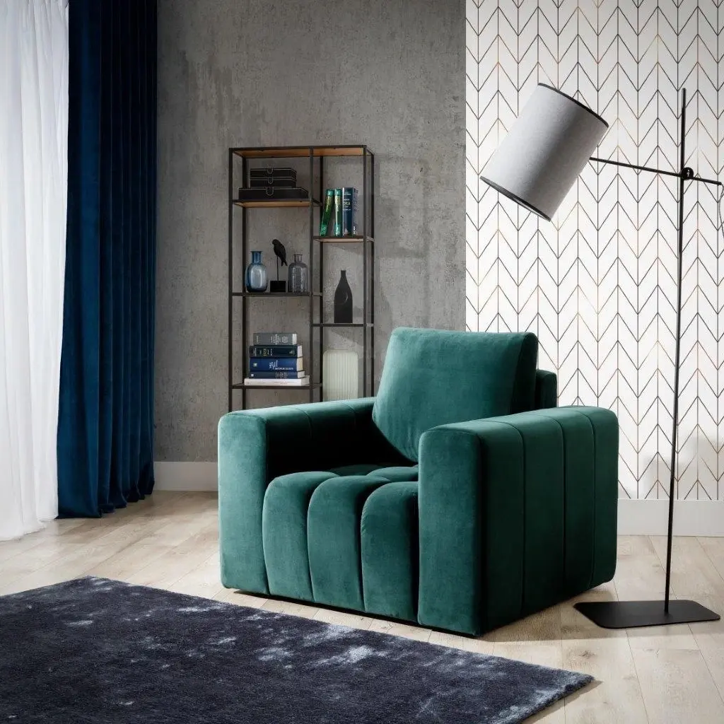 Welurowy fotel Lazaro w kolorze butelkowej zieleni jako wygodne miejsce do relaksu w salonie. Grube podłokietniki i głębokie siedzisko podkreślają komfort, obok stoi lampa podłogowa i dywan.