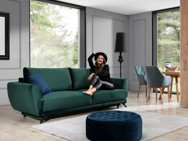 Elegancki pokój z butelkowozieloną rozkładaną kanapą Megis Riviera 38, granatową pufą Monty Monolith 77, lampą stojącą i oknami z widokiem na ogród.