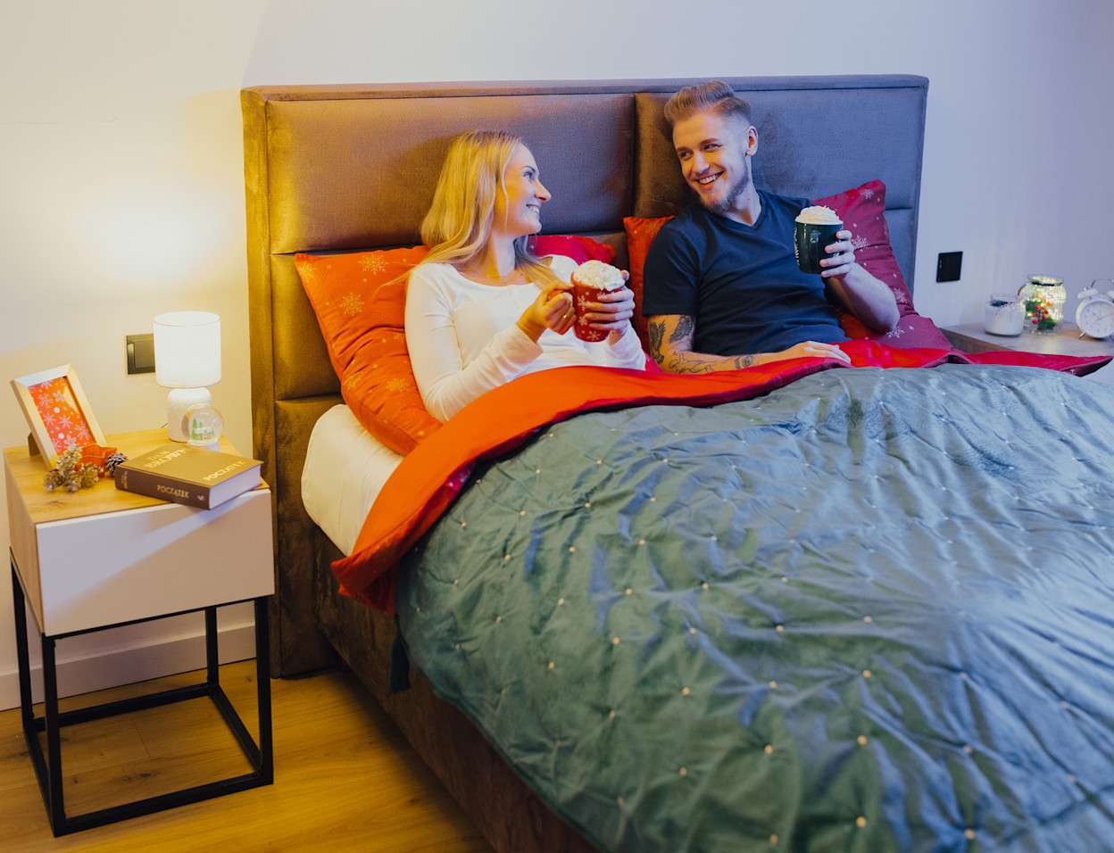 Łóżko Safiro w tkaninie Nube 05 w przytulnej sypialni, z wysokim tapicerowanym zagłówkiem i ciepłym oświetleniem. Para odpoczywa pod miękką kołdrą, co podkreśla komfort i domowy klimat wnętrza.