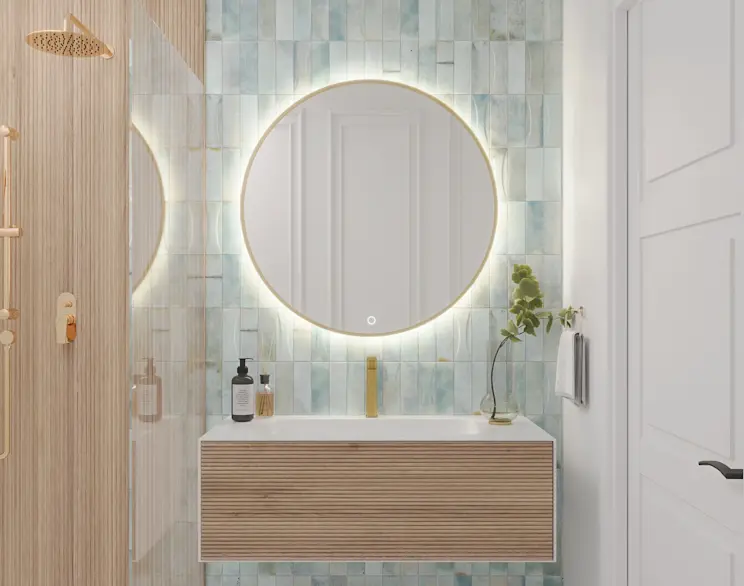 Okrągłe lustro LED Gerbinie w złotej ramie, zawieszone nad umywalką w nowoczesnej łazience. Widać równomierne podświetlenie dookoła tafli oraz dotykowy włącznik u dołu, a tło tworzą jasnoniebieskie płytki i drewniana szafka pod umywalką.