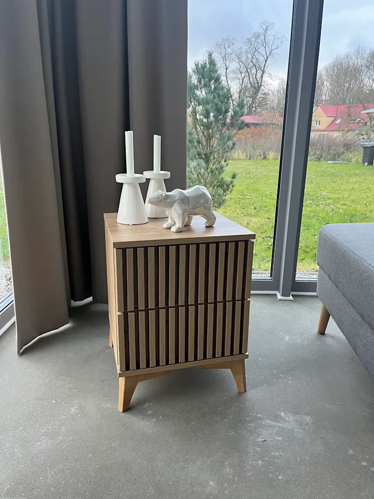 Szafka nocna Entsian z pionowymi lamelami, stojąca przy oknie z dekoracjami w postaci świeczników i porcelanowego niedźwiedzia.