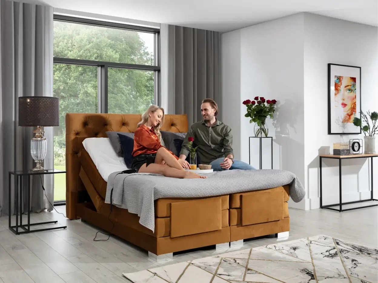 Regulowane elektrycznie łóżko Clover Monolith 48 w kolorze miodowym, w sypialni z dużym oknem; na łóżku para, obok stolik z lampą i dekoracje z różami.