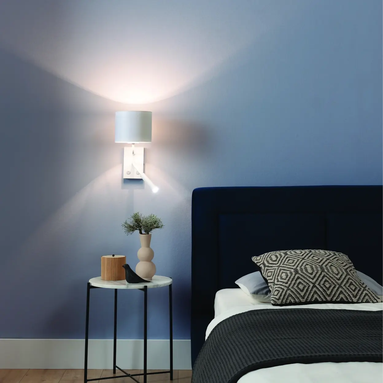 Biały kinkiet Vespillo na ścianie w sypialni, świecący miękkim światłem na niebieskiej ścianie. Obok stoi okrągły stolik z jasnym blatem i czarnymi nogami, z wazonem oraz dekoracjami. W kadrze widać także tapicerowane łóżko w ciemnym kolorze i poduszki.