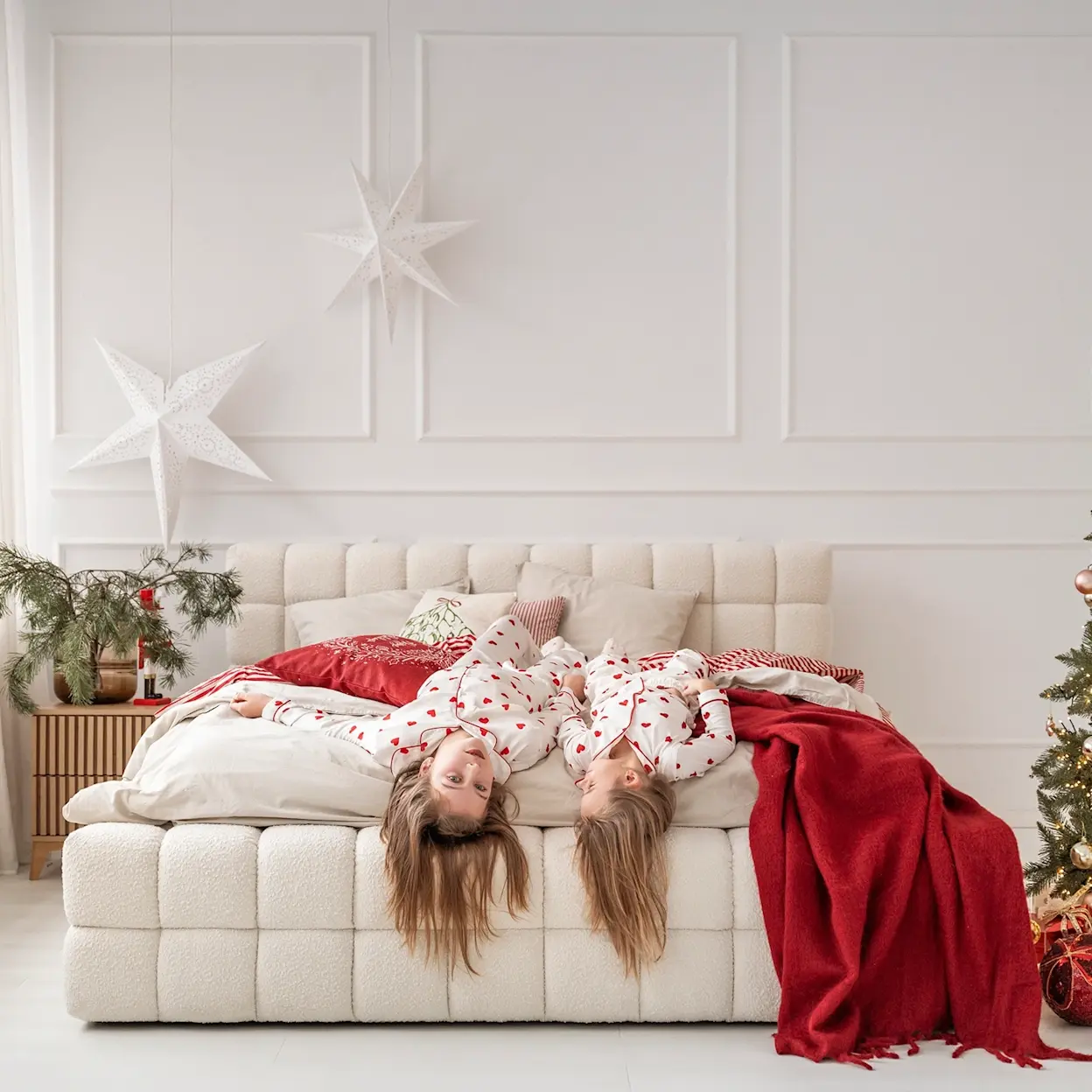 Świąteczna sypialnia z jasnym łóżkiem Dizzle tapicerowanym tkaniną boucle i wysokim zagłówkiem. Na łóżku leżą dzieci w piżamach, a czerwony koc, gwiazdy i choinka budują przytulny, rodzinny klimat.