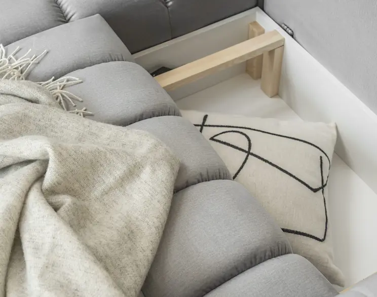 Szary narożnik z funkcją spania i pikowanym siedziskiem, pokazany z otwartym pojemnikiem na pościel. Praktyczne miejsce do przechowywania koców i poduszek, idealne do salonu w nowoczesnym stylu.