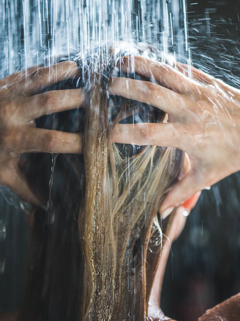 Mulher toma banho de costas lavando o cabelo.