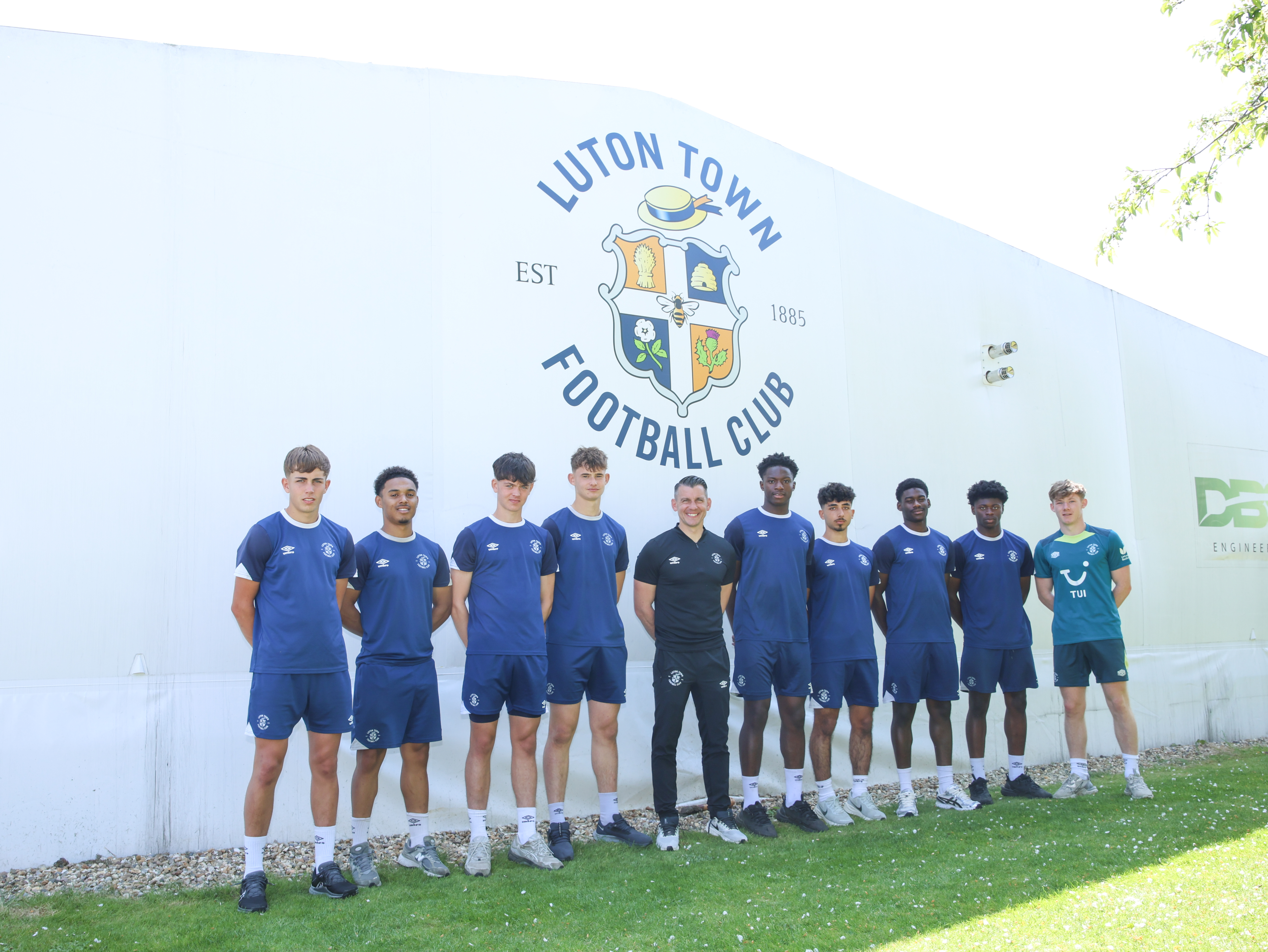 Manager Matt Bloomfield is pictured under the Luton Town club crest on the outside of the gym at The Brache with nine of the youth team players who have turned professional: (from left to right) - Charlie Trustram, Tate Xavier-Jones, Dylan Stitt, Lucas Thomas, Matt Bloomfield, Christian Chigozie, Zach Ioannides, Jamie Odegah, Benny Benagr, Sam Anderson.
