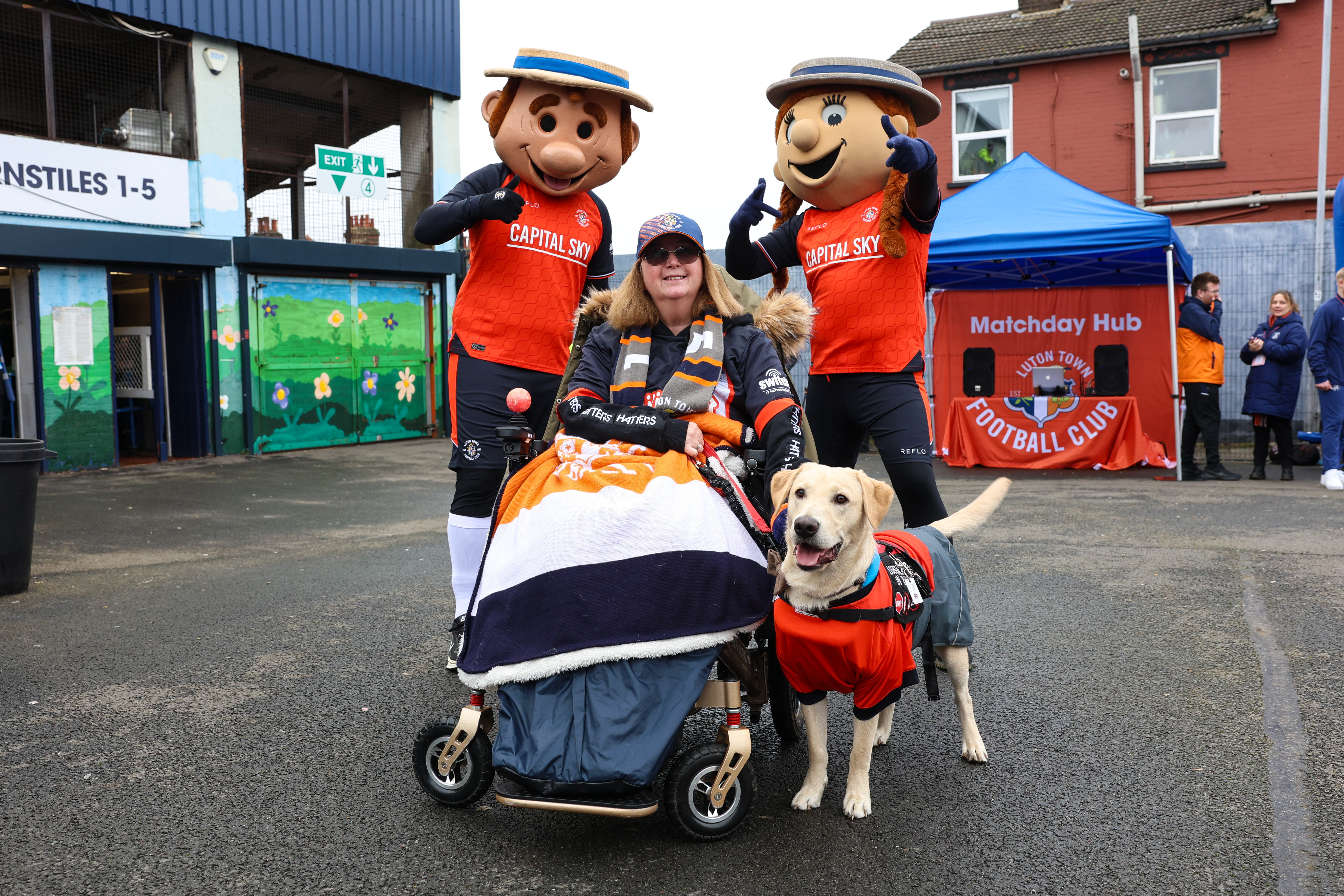 Tracy Camp and her assistance dog Milo pose with Happy Harry and Hatty outside Kenilworth Road.