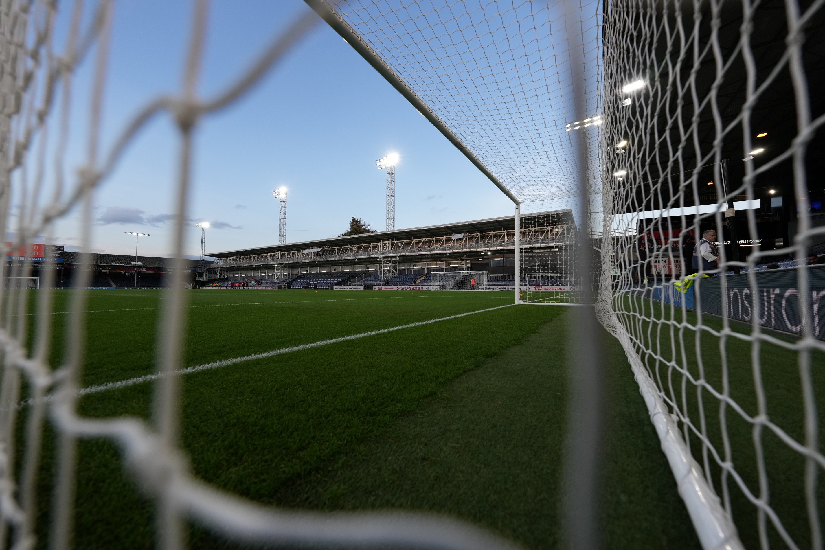 A view of the Bobbers stand at Kenilworth Road through the net at the Kenny End, with clear blue sky and the floodlights on.
