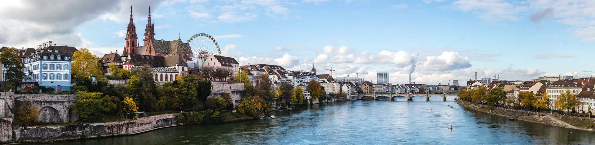 Panoramblick auf Basel mit dem Rhein, dem markanten Basler Münster und einem Riesenrad am Flussufer vor leicht bewölktem Himmel.