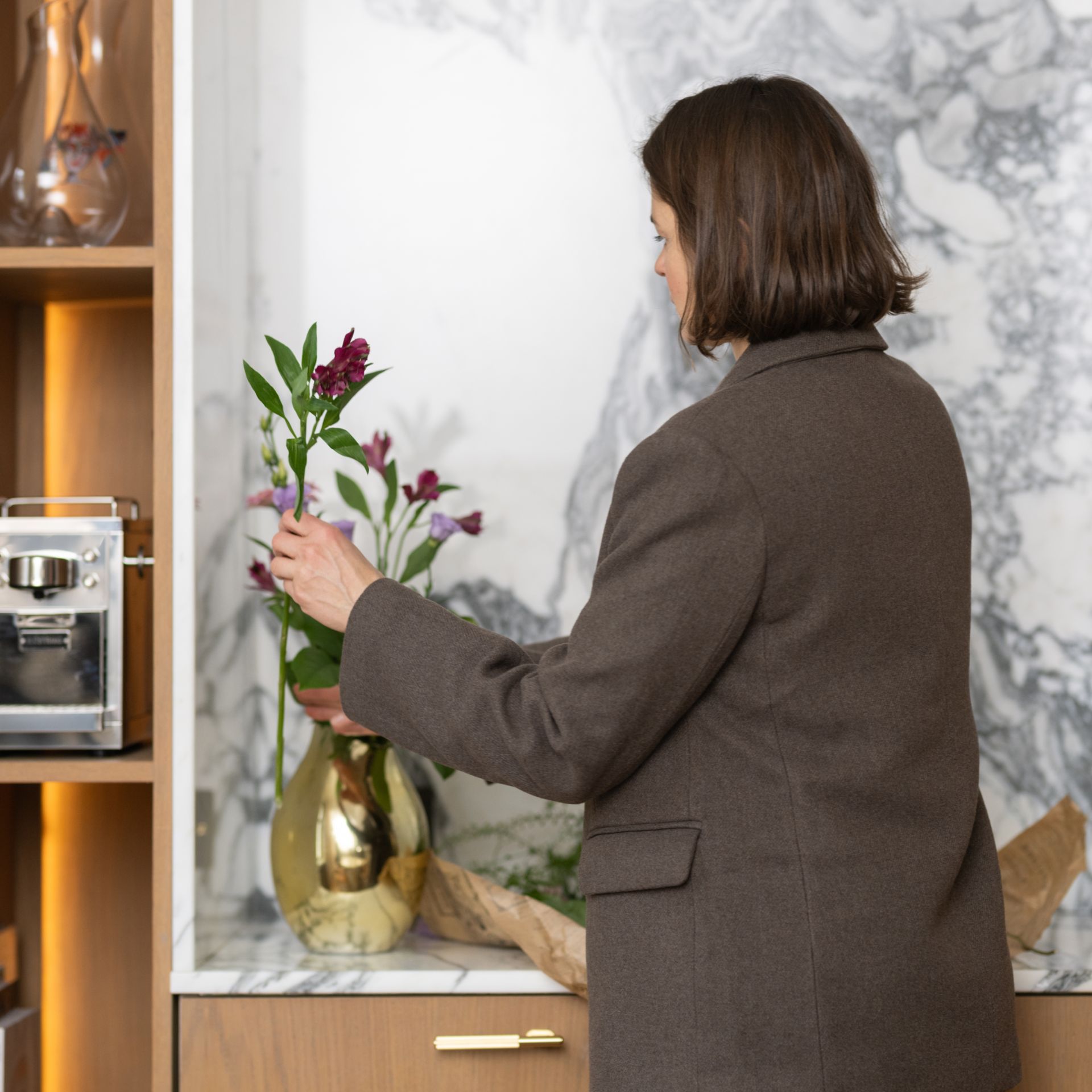 Kvinna i brun kavaj arrangerar blommor i en guldig vas på ett marmorbord.