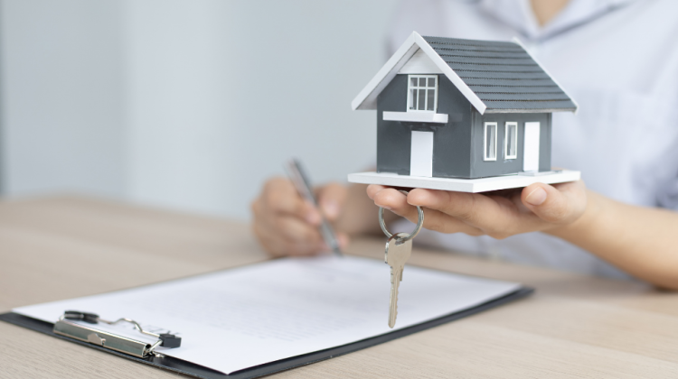person deciding whether to sell or rent out home, holding a small house and set of keys in their hand with a clipboard of paperwork on the table