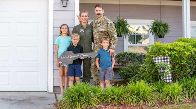 Dual military family holds a "Home Sweet Home" key sign in front of their new home. Was it better for them to purchase with a V Loan or Conventional Home Loan?