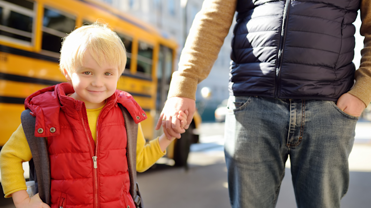 child holding the hand of an adult in front of a school bus, military family EFMP student at a new school