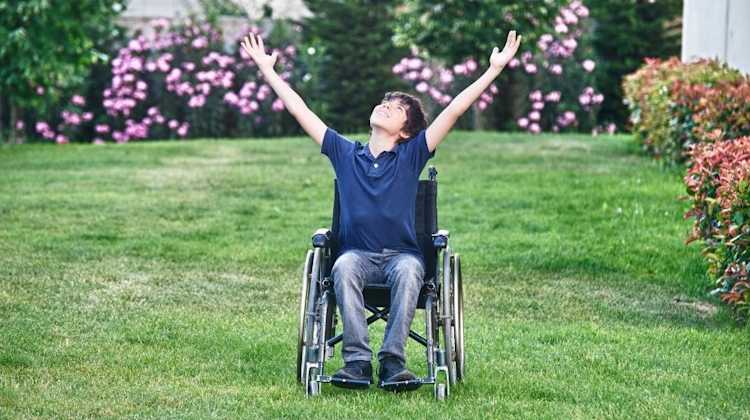 boy in wheelchair in a park, throwing his arms in the air in celebration