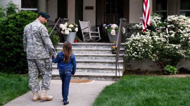 Army soldier holding child's hand in front of house