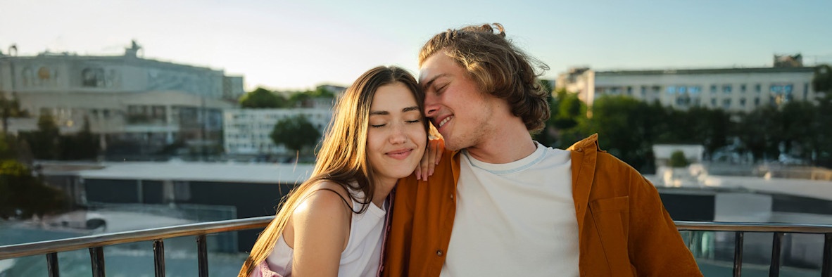 Ein Paar umarmt sich herzlich auf einem Dach bei Sonnenuntergang, beide mit gut gestyltem Haar.