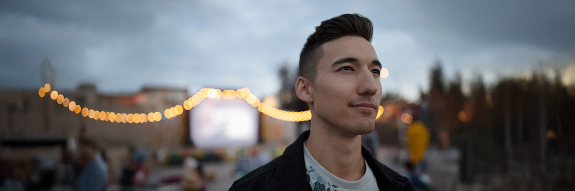 Ein junger Mann mit kurzen, dunklen Haaren posiert im Freien bei warmem, natürlichem Licht.