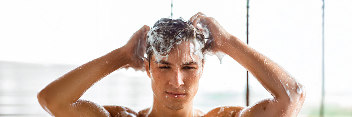 Ein Mann wäscht sich unter der Dusche die Haare, wobei sein Kopf von reichlich Schaum und Seifenblasen bedeckt ist.