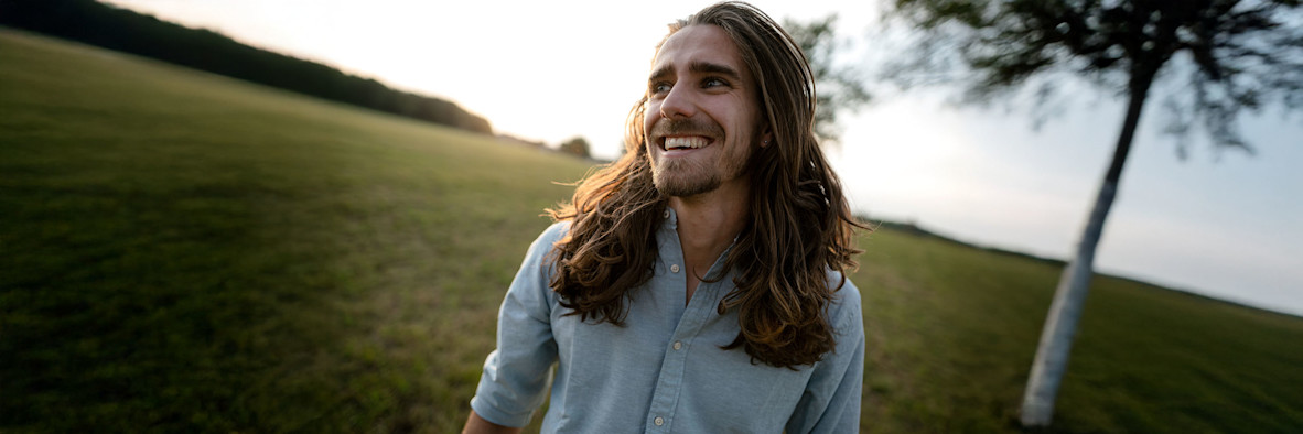 Ein Mann mit langen, natürlichen Haaren steht in einer offenen Landschaft, während der Wind durch sein Haar weht.