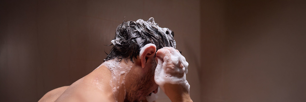 Ein Mann wäscht sich unter der Dusche die Haare und massiert dabei die Kopfhaut, während Shampoo aufschäumt.