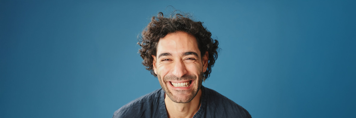 Ein lächelnder junger Mann mit natürlichen lockigen Haaren steht vor einem blauen Hintergrund.