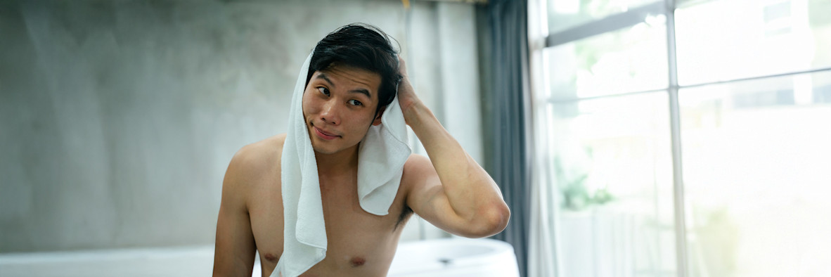 Ein dunkelhaariger Mann wickelt nach dem Waschen seine Haare in ein weißes Handtuch, im Spiegel des Badezimmers reflektiert.