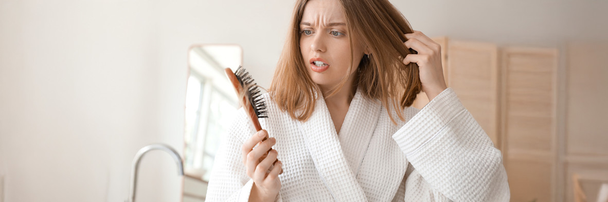 Eine Frau in einem weißen Bademantel bürstet sorgfältig ihr Haar und betrachtet es aufmerksam.