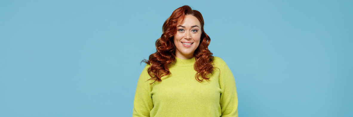 Eine fröhliche rothaarige Frau in einem gelbgrünen Oberteil lächelt vor einem passenden Hintergrund.