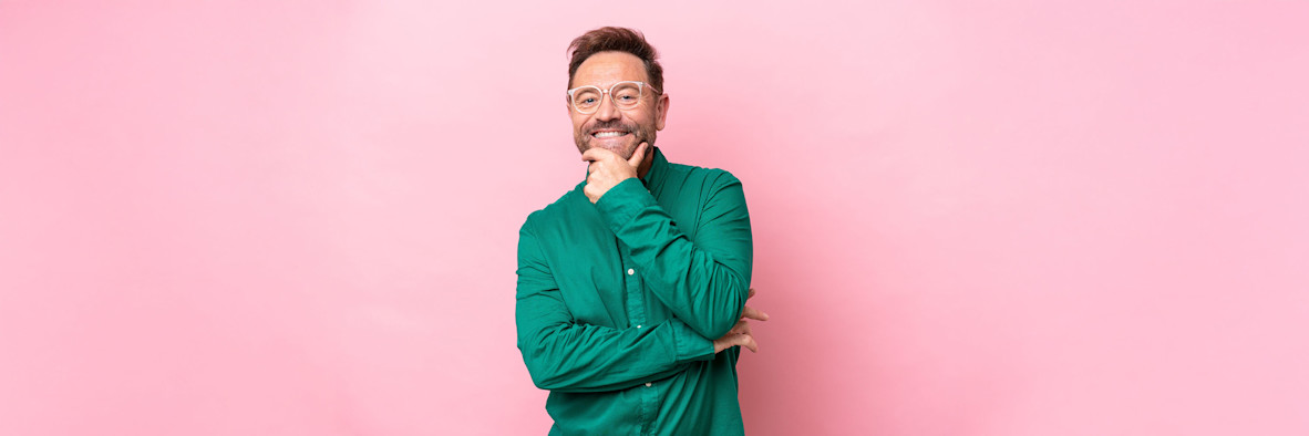 Ein bärtiger Mann in einem grünen Hemd lächelt breit vor einem rosafarbenen Studiohintergrund.