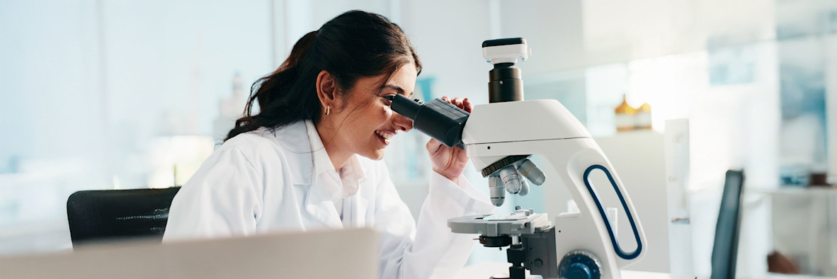 Eine Frau in einem weißen Laborkittel untersucht in einer klinischen Umgebung eine Probe unter einem Mikroskop.