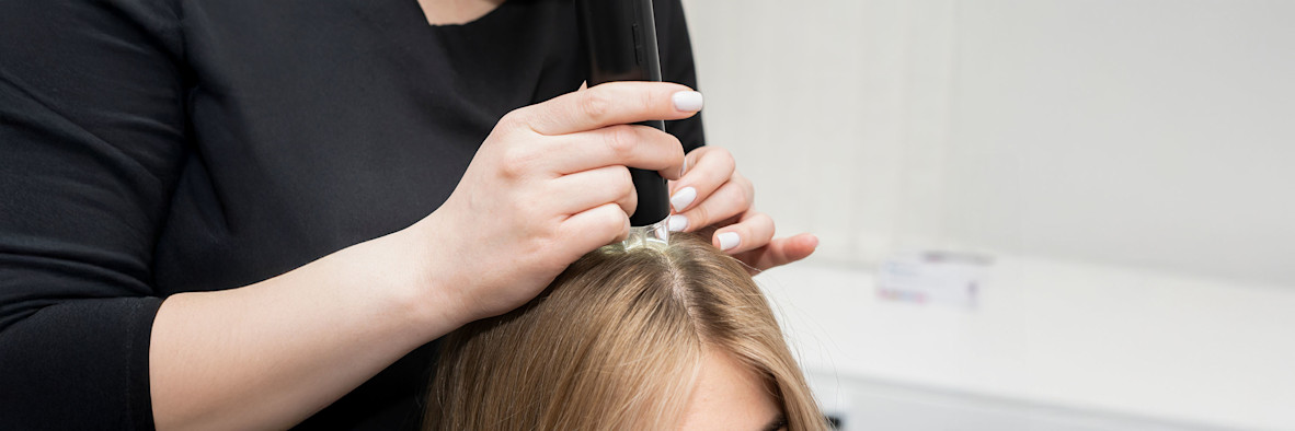 Eine Friseurin trägt ein Pflegeprodukt mit einem Applikator direkt auf den Scheitel einer blonden Frau auf.