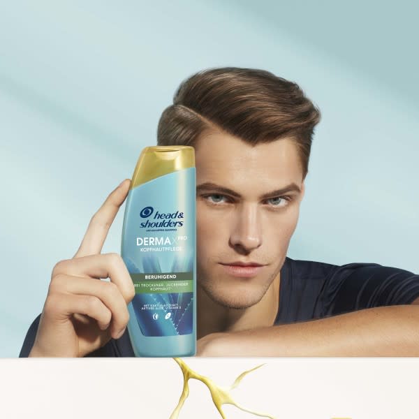 Ein Mann, der seinen Arm auf eine rissige Mauer lehnt und in der anderen Hand ein H&S DERMAXPRO Shampoo hält, welches auf der Mauer steht.
