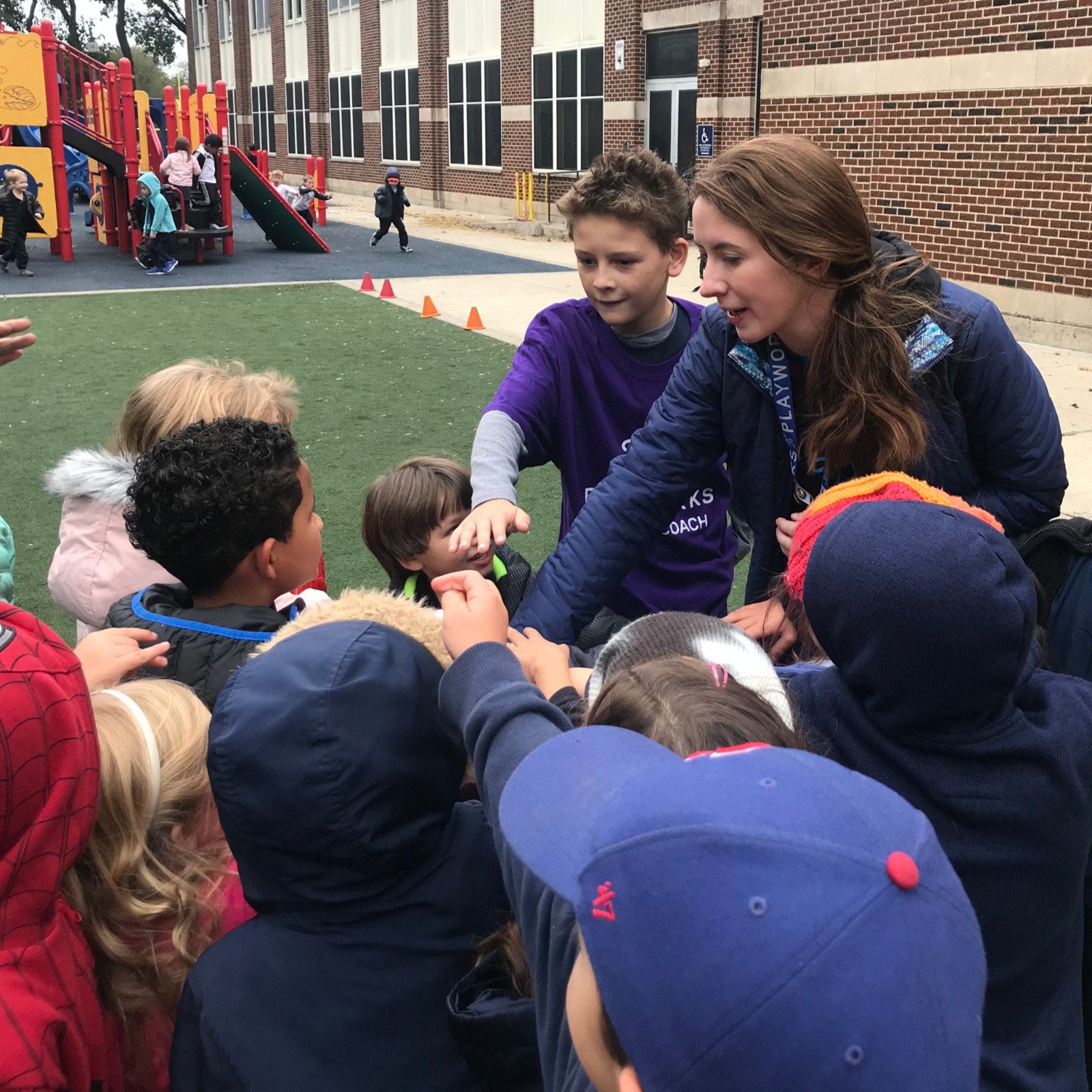 A fifth-grade junior coach named Jackson works alongside Ovitt to explain the rules of Sharks and Minnows to a group of students during recess. (Emily Tate / EdSurge)
