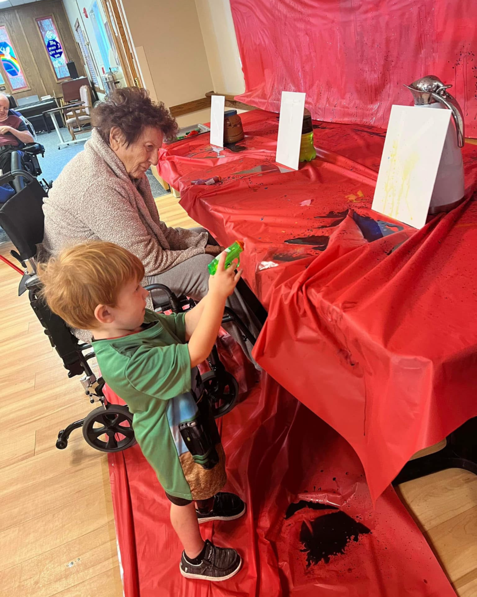 A child at Tiny Images paints a canvas using a squirt gun while a senior keeps watch. Photo courtesy of Tiny Images.
