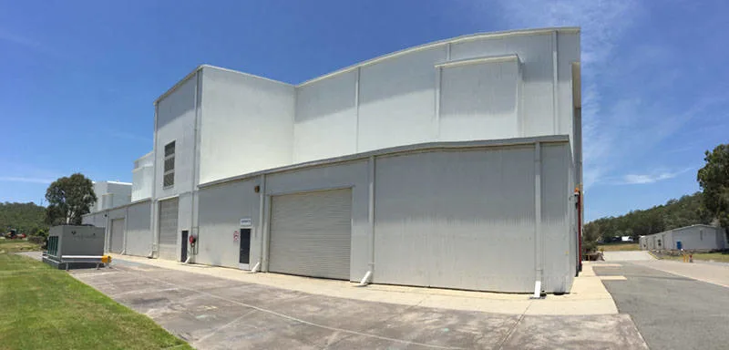 A large white industrial building with multiple garage doors and vents, set under a clear blue sky. There is a grassy area and trees nearby, and a smaller building is visible in the background.