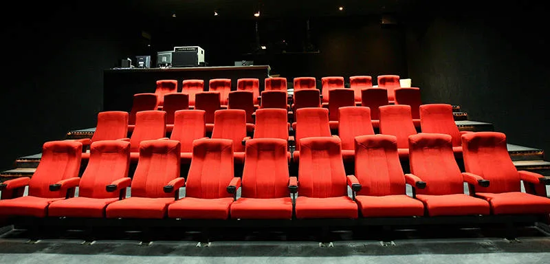 Rows of empty red seats in a small, modern movie theater, with a projection booth visible in the back. The room is dimly lit and has dark walls.