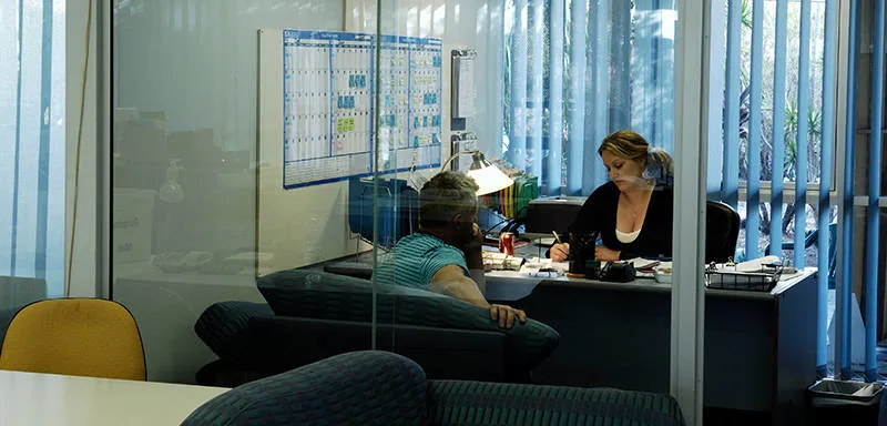 Two people sit across from each other at a desk in an office with glass walls. One person writes on paper while the other listens. A large wall calendar and desk lamp are visible, along with sunlight filtering through blinds.